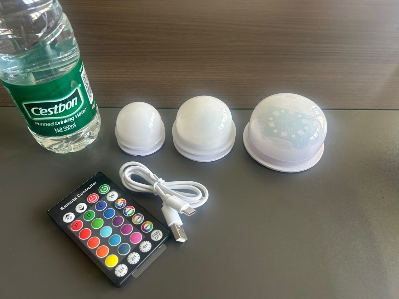 Three compact rechargeable LED modules beside a 350 ml water bottle, remote control and USB-C charging cable